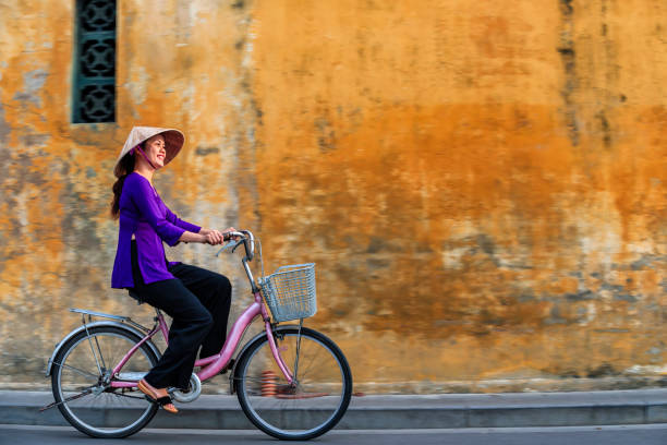 Vietnamese woman riding a bicycle, old town in Hoi An city, Vietnam Vietnamese woman riding a bicycle in old town in Hoi An city, Vietnam. Hoi An is situated on the east coast of Vietnam. Its old town is a UNESCO World Heritage Site because of its historical buildings. ao dai stock pictures, royalty-free photos & images
