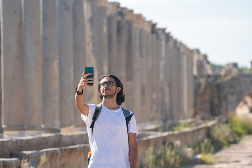 Photo of travelling young adult man with backpack in antique city of Perge, Antalya, Turkey.  He is wearing a white t-shirt and taking selfie via smartphone. Shot under daylight with a full frame mirrorless camera and a tele photo lens.