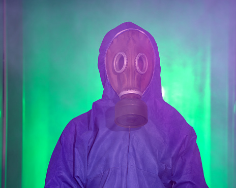 Portrait of man wearing a hazmat suit and old fashioned military gas mask in fog. The fog is colored in green. Shot in studio with a full frame mirrorless camera.