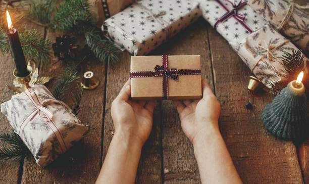 istock Christmas rustic gift. Hands holding stylish christmas gift wrapped in craft paper on rustic wooden table with candles, scissors, fir branches. Merry Christmas! Atmospheric moody image 1341338130