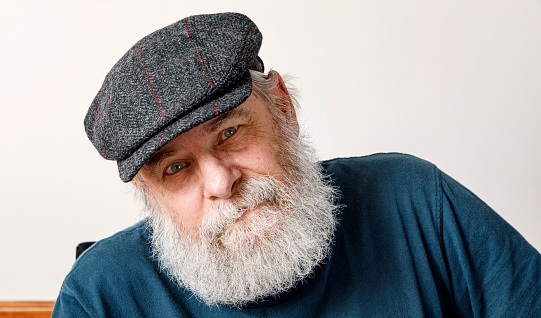 Senior adult man with a tangled gray beard wearing an Irish flat cap style hunting hat looking at the camera with a quizzical half smile.