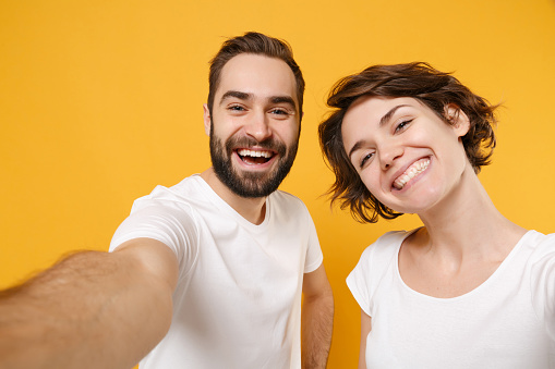 Close up of cheerful young couple friends bearded guy girl in white t-shirts isolated on yellow orange wall background. People lifestyle concept. Mock up copy space. Doing selfie shot on mobile phone