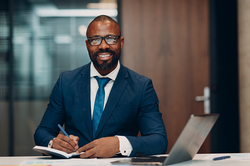 Retrato sonriente hombre de negocios afroamericano con traje azul se sienta a la mesa para reunirse en la oficina con cuaderno con bolígrafo y computadora portátil. photo
