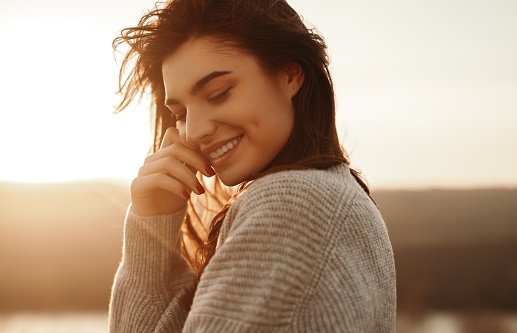 Side view of young long haired brunette in warm knitted sweater touching face and smiling while resting near lake at sunset time