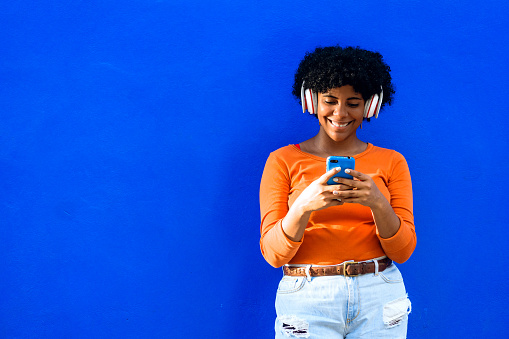Happy beautiful afro american woman with orange sweater andwhite headphones listening to music on a phone, music and technology concept.