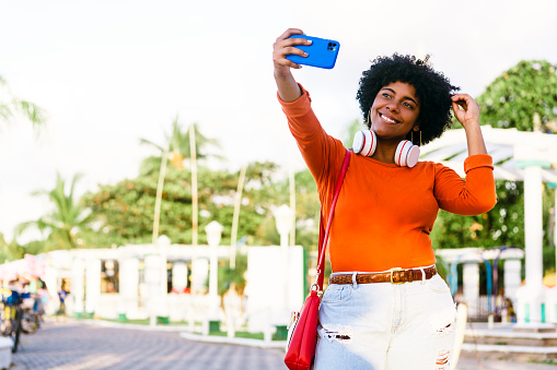 beautiful afro american woman with curly hair, wearing an orange shirt with headphones taking a picture with a mobile. technology and entertainment concept. honduras people.