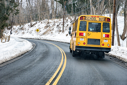 Car point of view looking through the windshield from a following car at a yellow school bus ahead sweeping left through an uphill curve just before an upcoming three way road intersection on a rural winter road on a rainy, snowy February day near Rochester, New York.