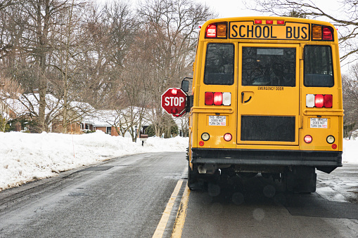 Car point of view looking through the windshield at the rear of a stopped yellow school bus with its automated stop signs and caution lights flashing while students enter or exit the vehicle. It's a rainy, snowy February day near Rochester, New York.