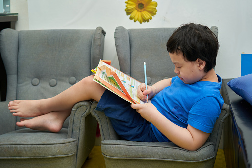Asian boy sitting on sofa with homework book and use a pencil to do the assigned homework at home