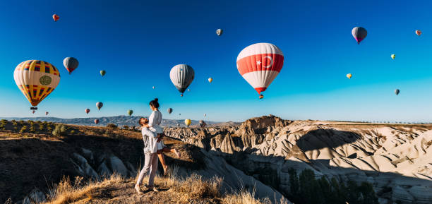 A happy young couple enjoys balloons during sunrise in Cappadocia, Turkey, panorama. A man and a woman on vacation in Turkey watch balloons. Honeymoon trip. Travel to the tourist places of Turkey stock photo