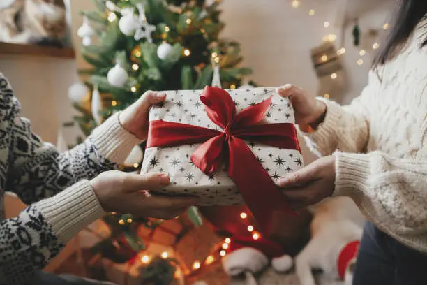 Couple exchanging christmas gift with red bow on background of christmas tree with lights. Stylish couple hands holding present with red ribbon close up in festive decorated room. Happy Holidays