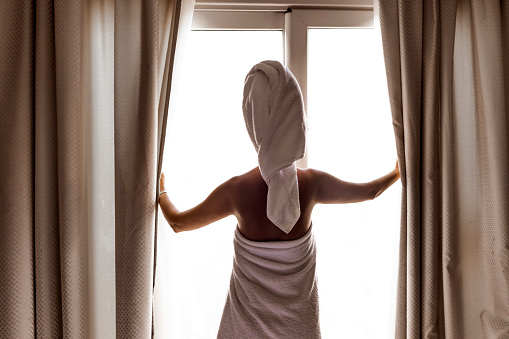 Rear view of a young woman wrapped in towel standing, looking through the window and opening patio doors.