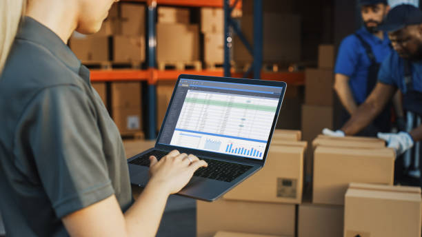 female manager using laptop computer to check inventory. in the background warehouse retail center with cardboard boxes, e-commerce online orders, food, medicine, products supply. over the shoulder - logistical imagens e fotografias de stock