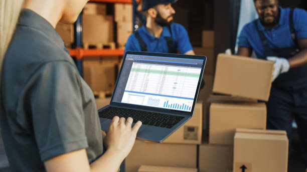 istock Female Manager Using Laptop Computer To Check Inventory. In the Background Warehouse Retail Center with Cardboard boxes, e-Commerce Online Orders, Food, Medicine, Products Supply. Over the Shoulder 1341393493