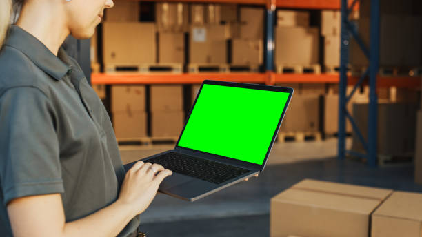 female manager using green screen chroma key laptop. in the background warehouse retail center with cardboard boxes, e-commerce online orders, food, medicine, products supply. over the shoulder - logistical imagens e fotografias de stock