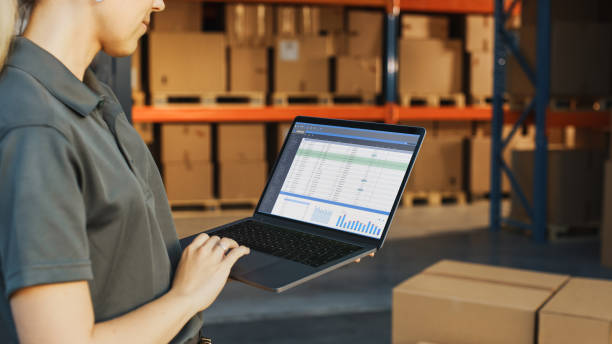 istock Female Manager Using Laptop Computer To Check Inventory. In the Background Warehouse Retail Center with Cardboard boxes, e-Commerce Online Orders, Food, Medicine, Products Supply. Over the Shoulder 1341393505