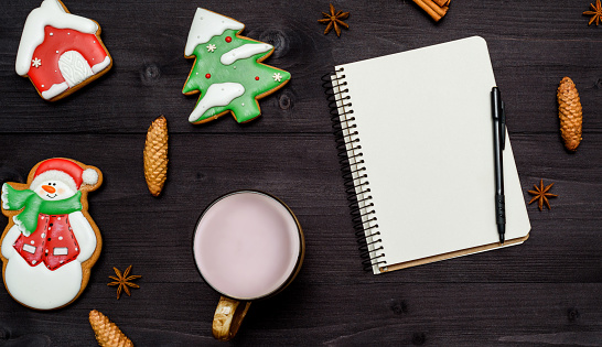 Open blank notebook, cup of hot cocoa and homemade gingerbread cookies on dark wooden desk. Top view, flat lay. Christmas background. New year resolutions, goals, free space
