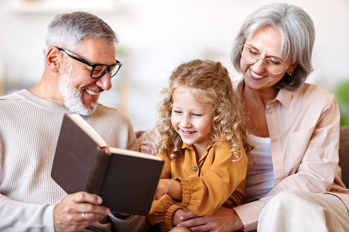 Cute little girl granddaughter smiling when reading book with senior grandparents while sitting together on sofa in living room, child enjoying leisure time with grandma and grandpa on weekend at home