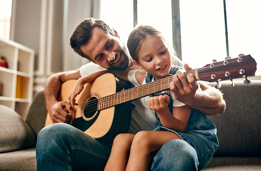 Happy dad teaches his cute daughter to play the guitar while sitting on the sofa in the living room at home. Happy Father's Day.