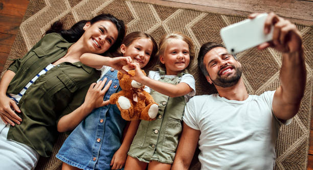 Family at home Dad, mom and two cute daughters lie on the carpet on the floor in the living room and take a selfie on a smartphone. Happy family have fun together. family at home stock pictures, royalty-free photos & images