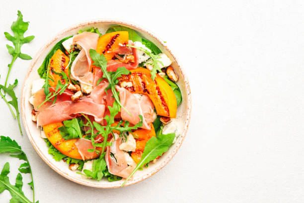 istock Salad bowl with grilled peach, prosciutto, goat cheese, walnuts and arugula on white background, top view, copy space 1341401620