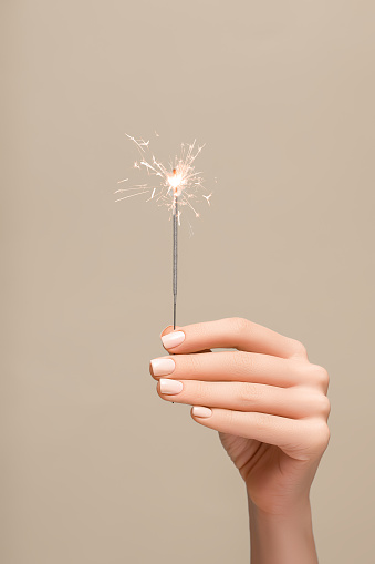 New Year lighted sparkler in female hand on beige background. Woman hold glowing holiday sparkling fireworks, shining fire flame. Christmas light.