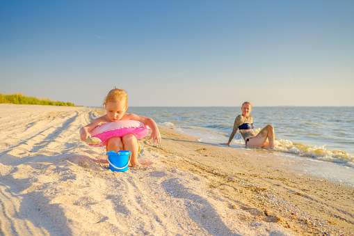 Little blonde girl plays in the sand on the sea beach. In the background, the baby's mom relaxes and sunbathes by the water. Family vacation at the sea.
