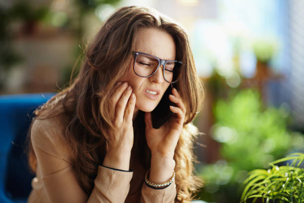 sad elegant housewife in house in sunny day talking on phone Green Home. sad elegant middle aged housewife with long wavy hair with toothache speaking on a smartphone in the modern house in sunny day. toothache stock pictures, royalty-free photos & images