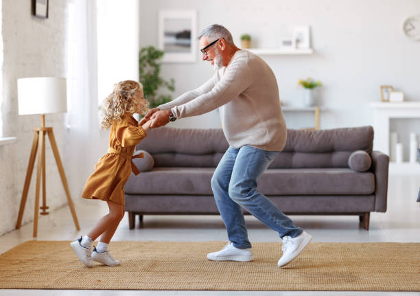 aktiver älterer großvater, der mit süßer kleiner enkelin im wohnzimmer zu hause tanzt - grandparent grandfather granddaughter little girls stock-fotos und bilder