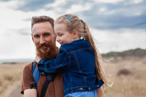 Father with daughter in hands standing on a hill with nature lanscape. Little blonde girl smiling and hugging with dad outdoor. Loving child embrace her daddy. Father's Day family greeting card.