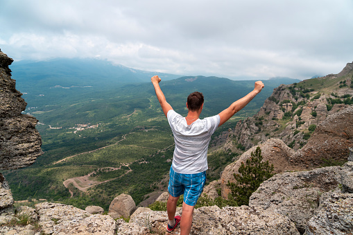 Young man raising hands up on top of mountain and looking to valley and peaks below. Healthy outdoors lifestyle concept. Concept of travel lifestyle and vacation in mountains