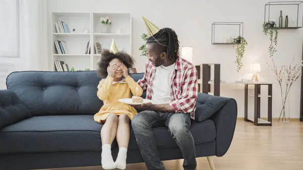 Caring african dad in party hat presenting birthday cake to little daughter, joy