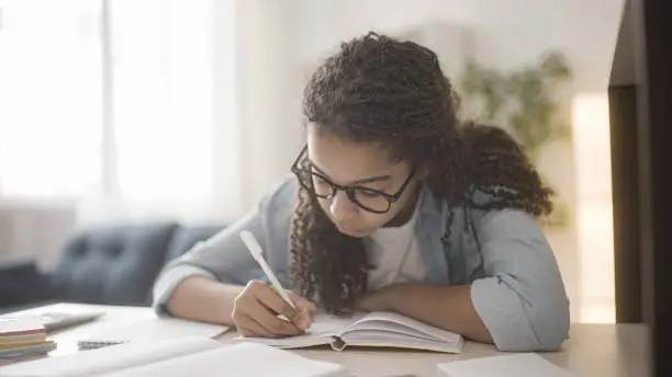 Teenage girl in glasses doing homework, writing in notebook, distance education