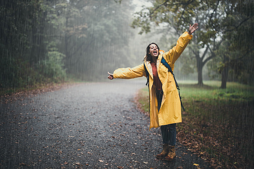 Young happy woman in yellow raincoat having fun while hitchhiking during rainy day in the forest. Copy space.