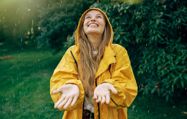 Image of a positive young blonde woman smiling wearing yellow raincoat during the rain in the park. Cheerful female enjoying the rain outdoors. Woman looking up and catching the rain drop with hands. stock photo