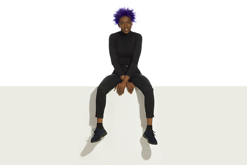 Young african american woman with short purple hair, wearing black clothes is sitting relaxed on a top of white banner. Full length studio shot.