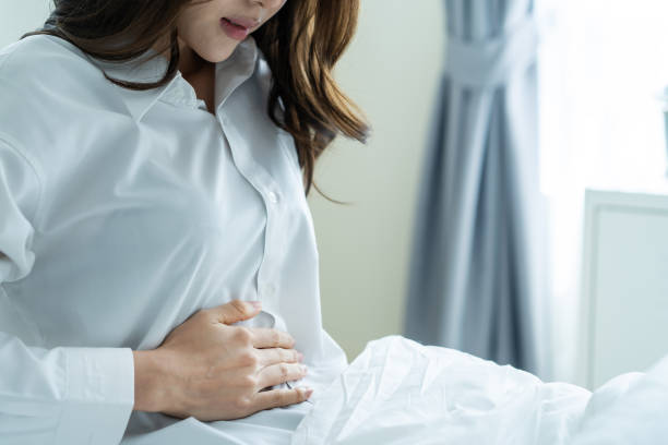 パジャマでアジアの美しい病気の女の子は寝室で睡眠から起きること。魅力的な若い女性は、ベッドで目を覚ました後、胃の痛みや生理を過ごし、朝に腹を抱いた後、悪いと痛みを感じます� - 腹痛 ストックフォトと画像