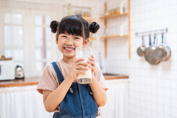 portrait of asian little cute kid holding a cup of milk in kitchen in house. young preschool child girl or daughter stay home with smiling face, feel happy enjoy drinking milk and then look at camera. - milk child drinking little girls imagens e fotografias de stock