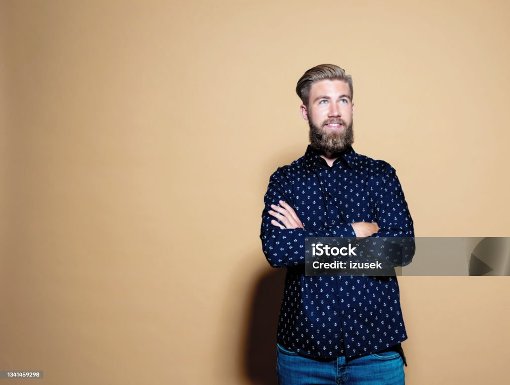 Confident entrepreneur standing over brown background Portrait of confident entrepreneur. Bearded young businessman standing with arms crossed against brown background, looking away. He is in casuals. Professional Occupation Stock Photo