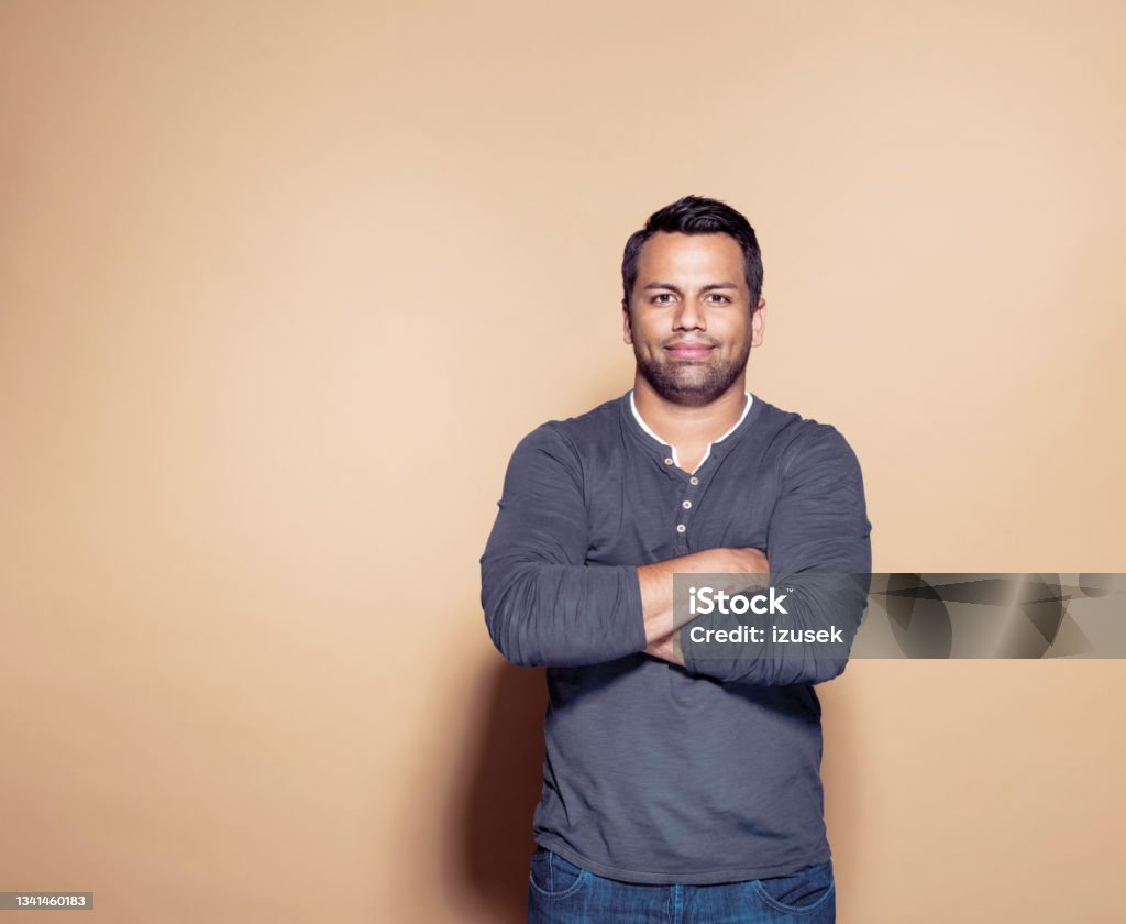 Confident man over brown colored background Portrait of entrepreneur looking at camera. Confident mid adult man standing against brown background with arms crossed, looking at camera. Men Stock Photo