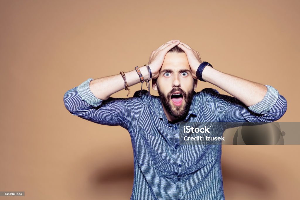 Portrait of shoced bearded man Portrait of terrified entrepreneur staring at camera with mouth open. Confident businessman standing against brown background. He is in casuals. Shock Stock Photo