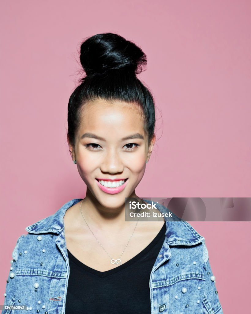 Smiling young woman against pink background Portrait of confident asian young woman. Female entrepreneur wearing oversized denim jacket standing against pink background, looking at camera. Hair Bun Stock Photo