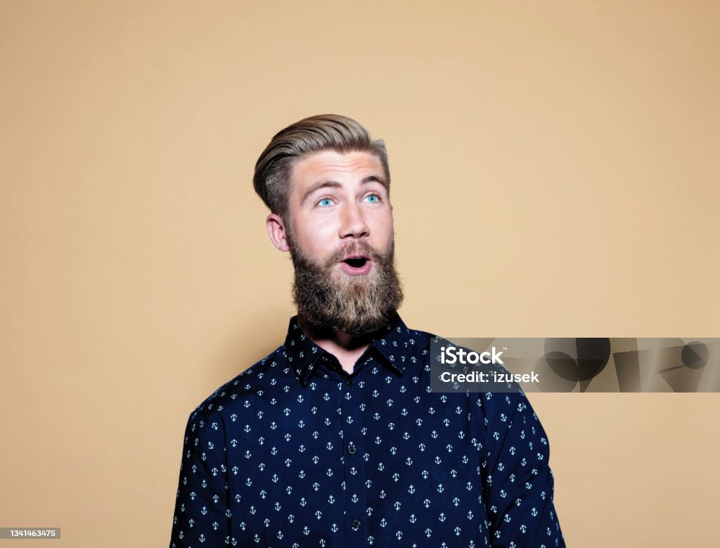 Surprised young man standing over brown background Portrait of happy entrepreneur. Bearded young businessman with toothy smile standing against brown background, looking away. He is in casuals. Beard Stock Photo