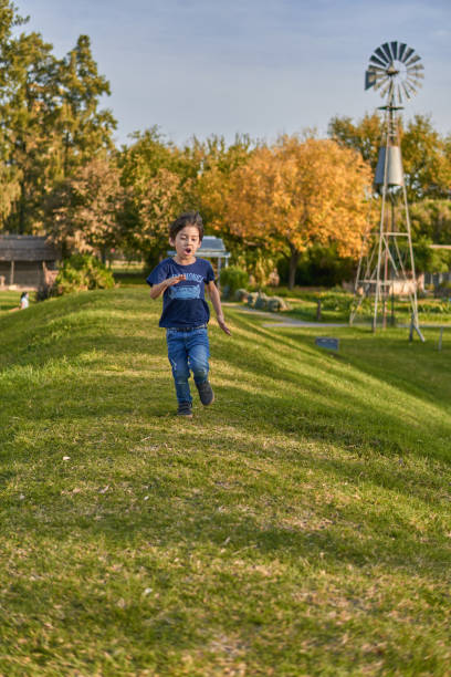 pequeño niño moreno corriendo por una colina y detrás de él hay un molino de viento en otoño en el bosque. vertical - run of the mill fotografías e imágenes de stock