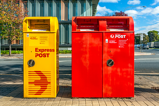 Adelaide, Australia - May 12, 2021: Corner Greenhill Rd & Hamilton Blvd, Wayville: Front view Australia Post Street Yellow & Red Posting Boxes on busy intersection with modern office building & traffic in the background