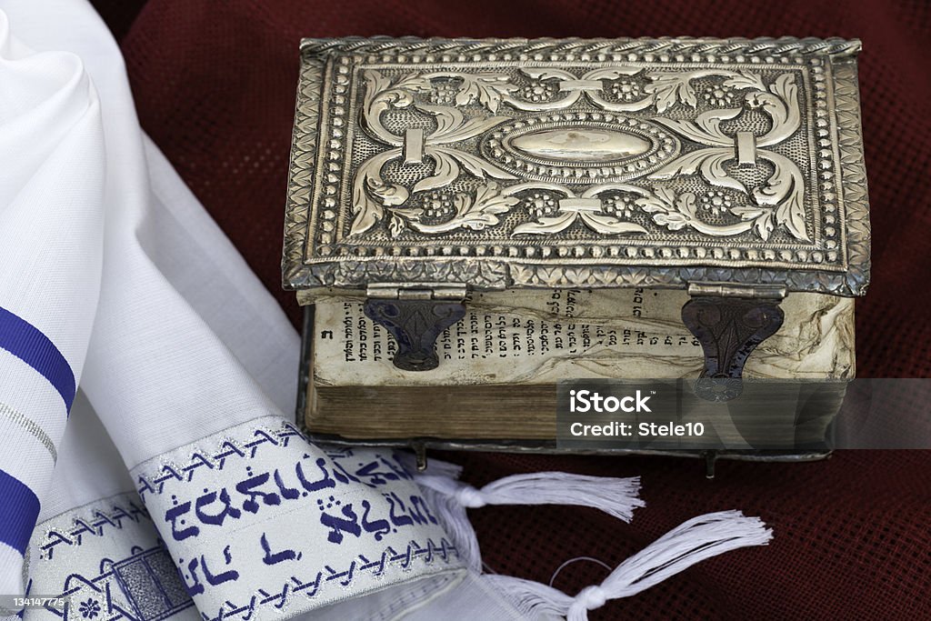 Antique Prayer Book and Tallit On A Purple Background An antique jewish prayer book, dated 1772, italian publisher, in a precious silver, embossed, baroque cover, and a tallit on the left, laying on a purple woolen background. Ancient Stock Photo