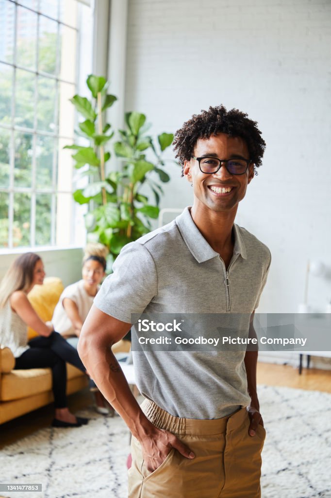 Young male designer smiling while standing in a modern office Portrait of a smiling young African American designer standing in an office with two colleagues working on a sofa in the background Millennial Generation Stock Photo