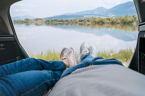Two pairs of jeans dressed legs in white sneakers. Couple relaxing in camper trunk and enjoying mountain autumnal lake views. Cozy early autumn couple auto traveling concept image.