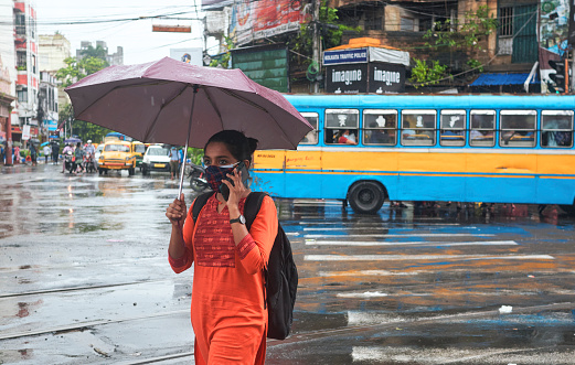 Hazra, Kolkata, 09/20/2021: A young office going girl wearing double protective face mask (due to coronavirus pandemic) unmindfully crossing street in a monsoon day.