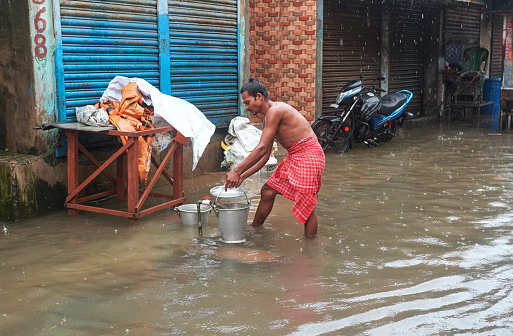 Kalighat, Kolkata, 09/20/2021: A shirtless young person collecting fresh / drinking water from street tap, unhygienically surrounded by dirty rain water. The street is heavily flooded caused by heavy overnight rain. All the nearby shops are seen closed.